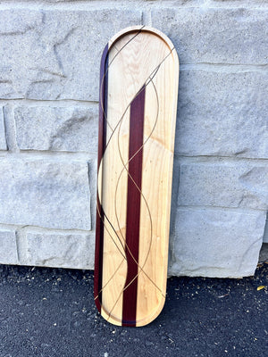 Long maple serving tray with purpleheart stripes and swirling walnut inlay lines, standing against a stone wall